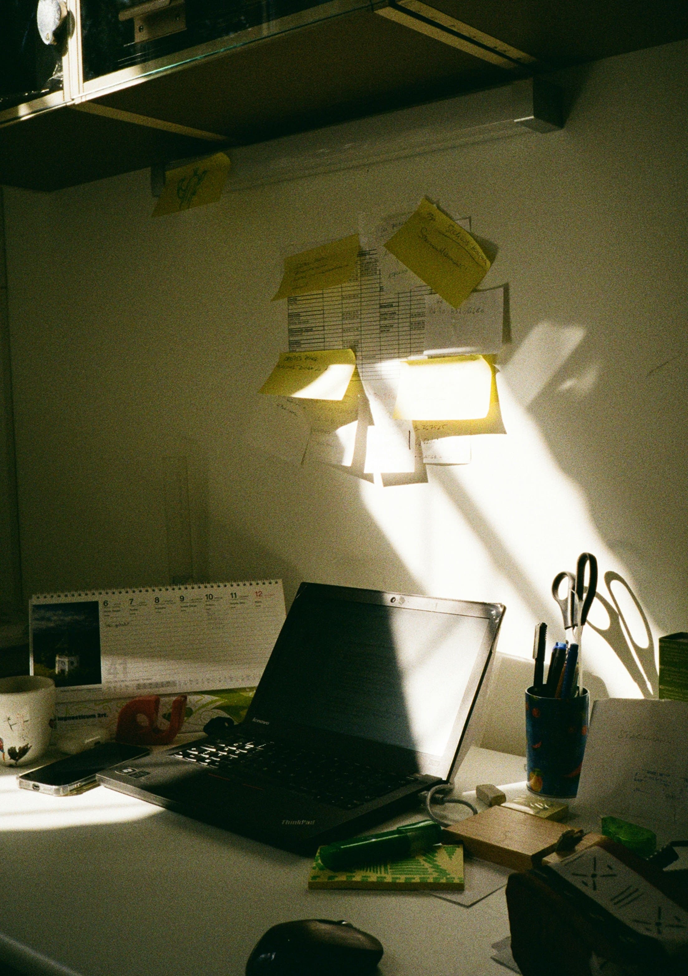 White desk with an open laptop, desk calendar, smartphone, mug, pen holder, yellow sticky notes on the wall, and sunlight across the surface